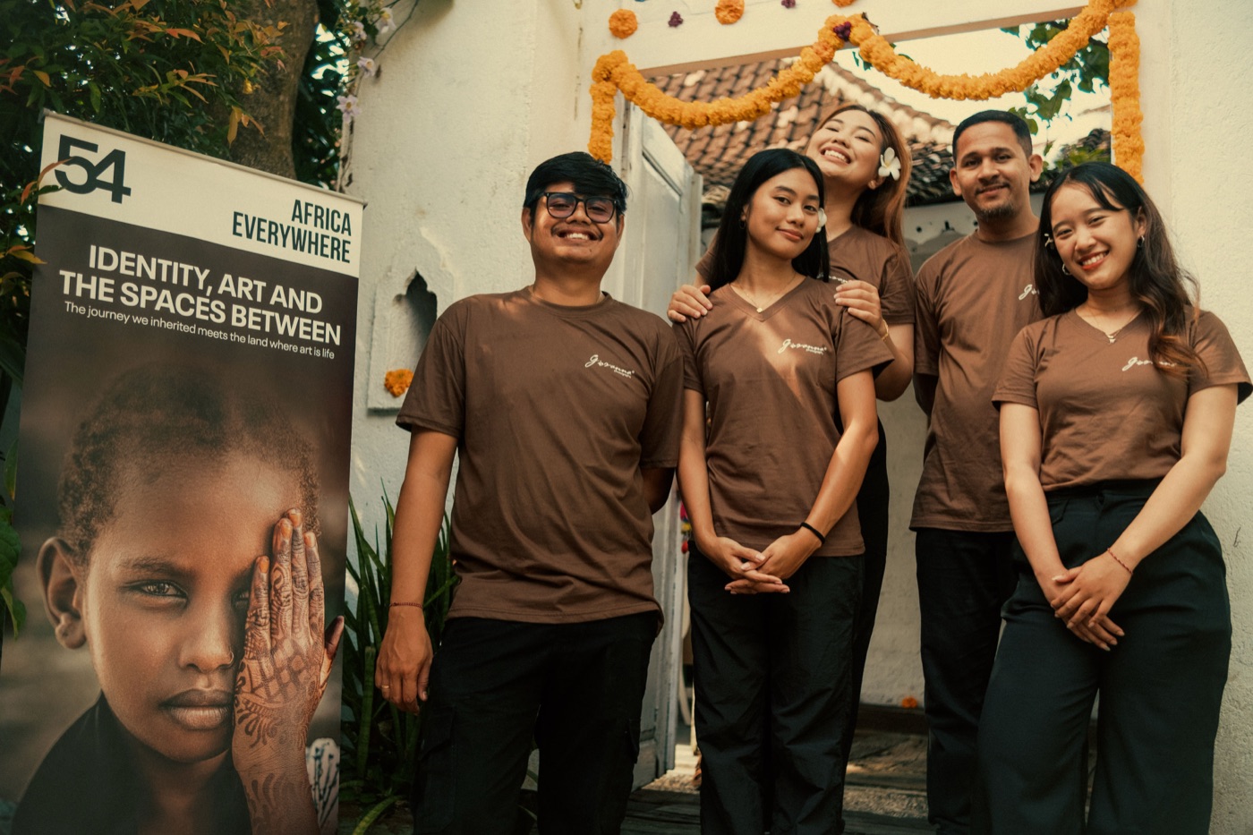 The Issue 13 banner — &ldquo;Identity, Art and the Spaces Between&rdquo; — beside the Giovana team under marigold garlands