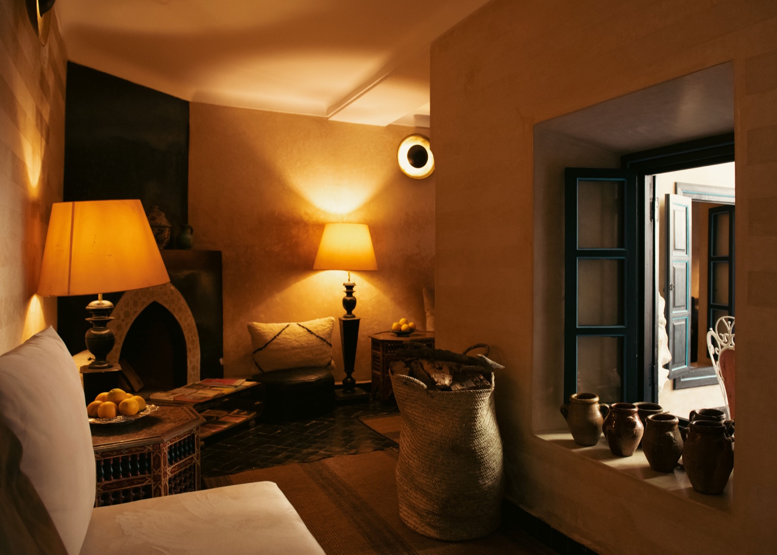 Fireside corner in warm tadelakt — lamplight, a carved side table and a window onto the patio